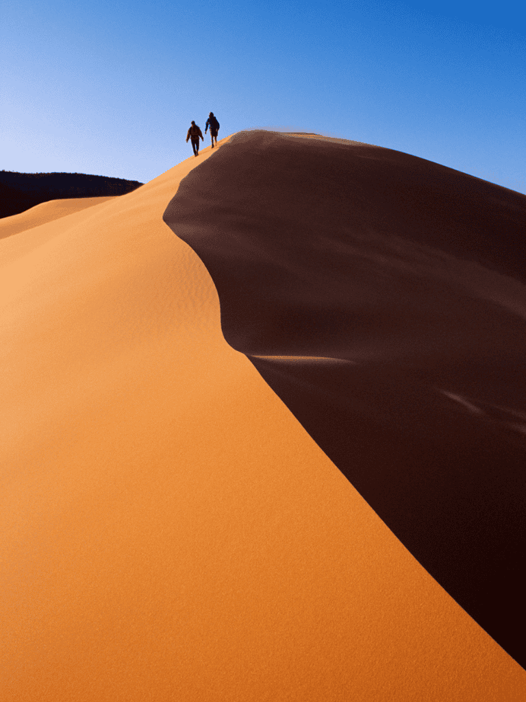 Vast desert landscape with two hikers on a sand dune under a clear blue sky.