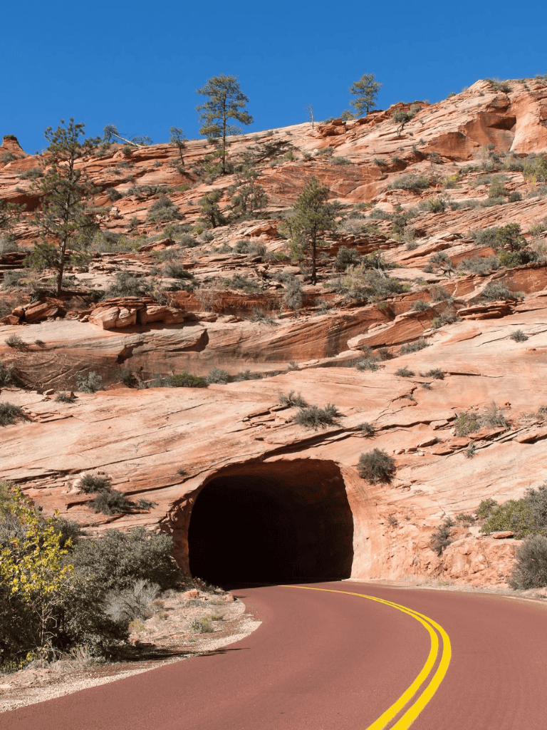 Vibrant desert landscape with tunnel entrance through red rock canyon, scenic highway, and sparse desert shrubs.