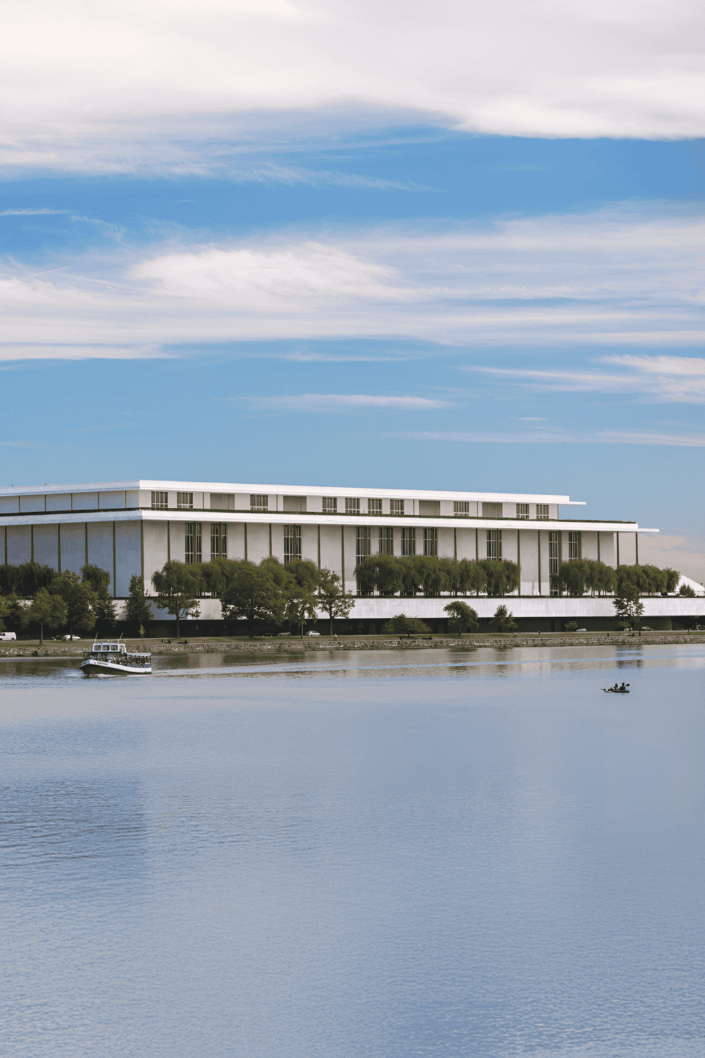 Modern waterfront building with lush trees and a boat on calm water under a blue Sky.