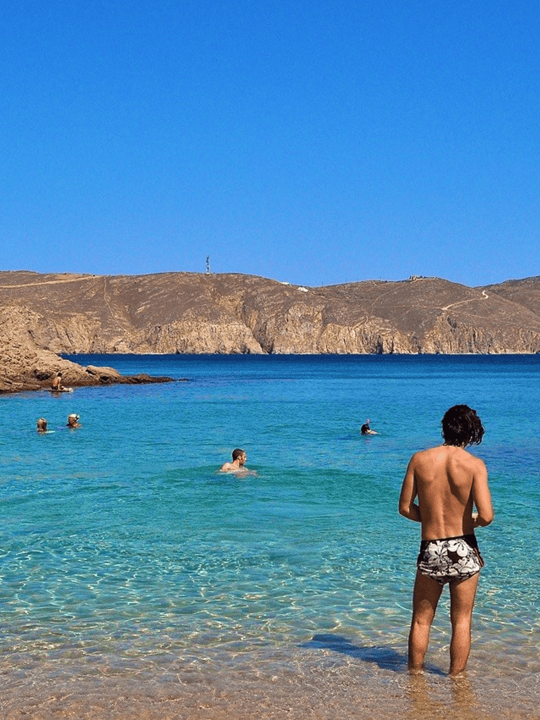 Calm blue sea with people swimming and exploring, sandy coastline, and rugged hills in the background.