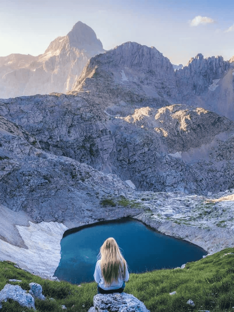 Serene woman sitting by mountain lake in alpine landscape, stunning view of rugged peaks and clear waters.