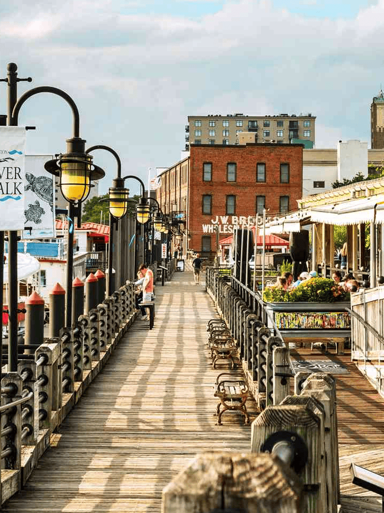 Scenic waterfront boardwalk with outdoor dining, historic buildings, and city skyline views.