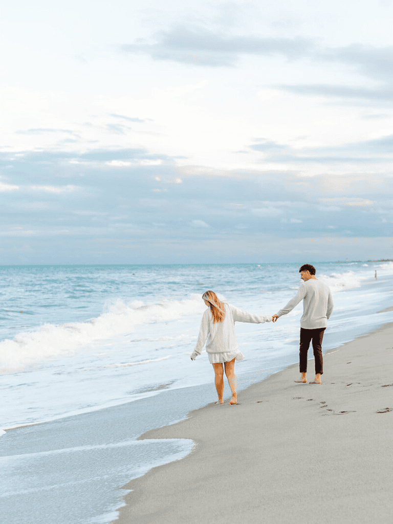 Seaside couple walking hand-in-hand on sandy beach during cloudy weather.