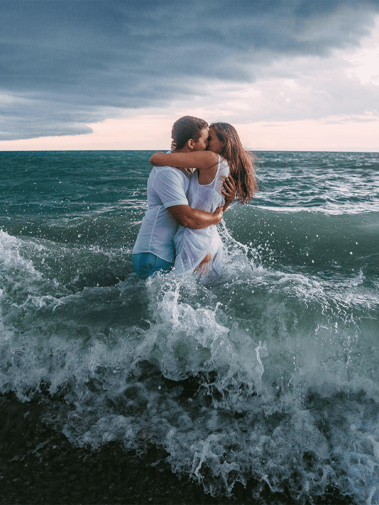 Romantic couple kissing in ocean waves with stormy sky, love, beach, and water theme.