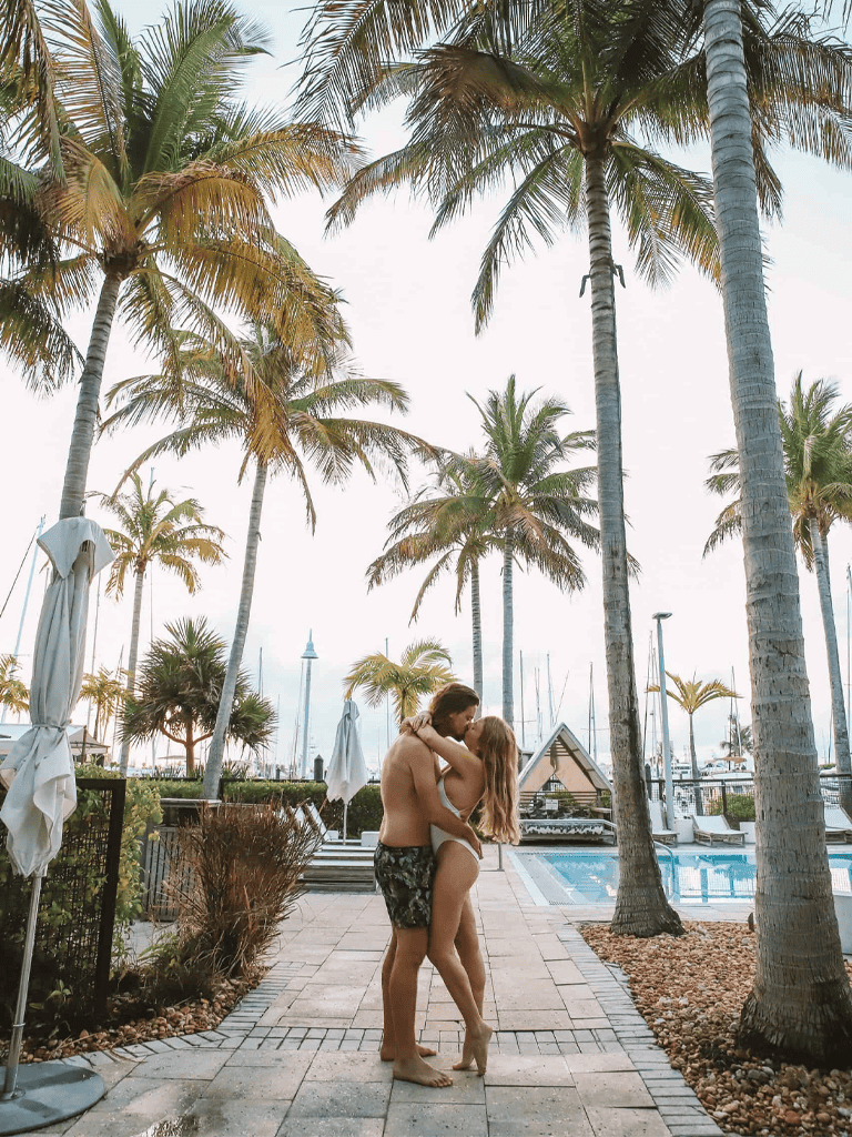 Romantic couple embracing by poolside under tall palm trees, tropical destination, summer vacation, paradise getaway, beach resort.