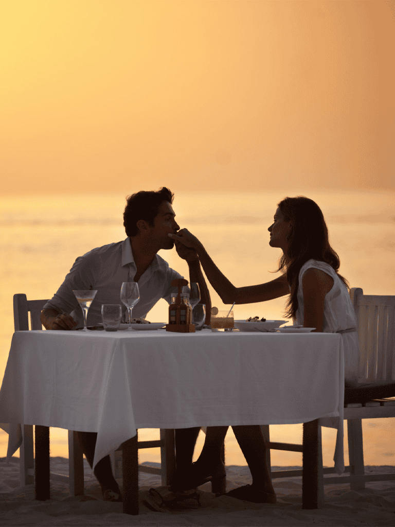 Enjoying romantic dinner on beach at sunset, couples celebrating love by the ocean.