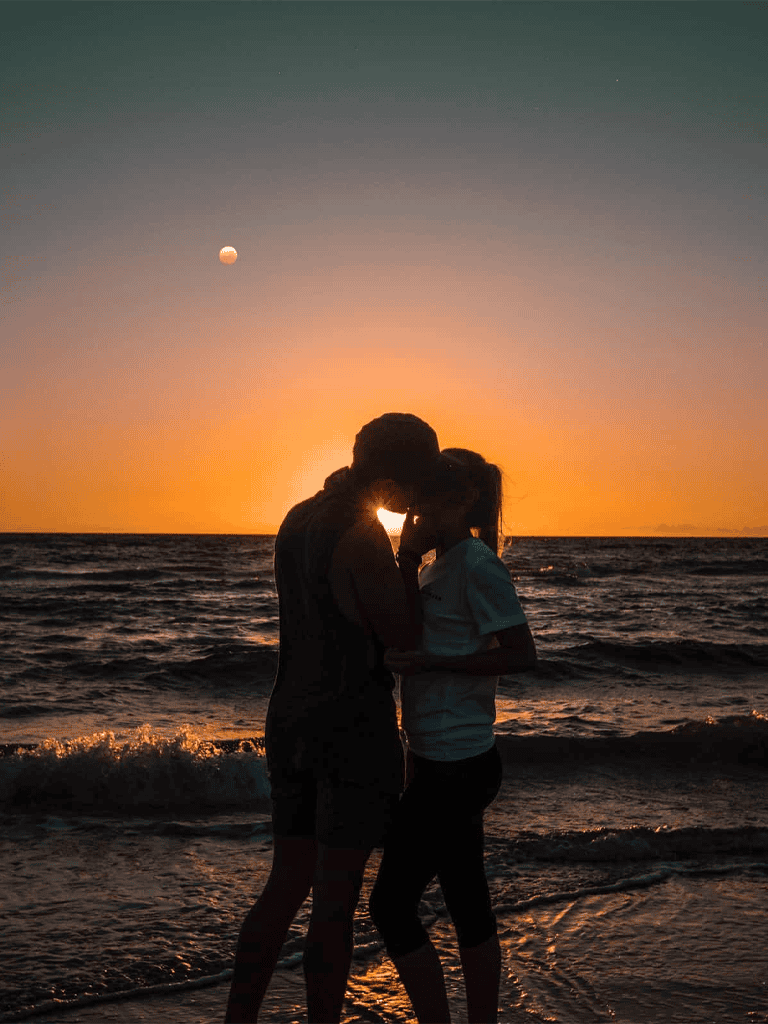 Silhouette of a couple kissing on a beach during sunset with ocean waves and a moon in the sky.