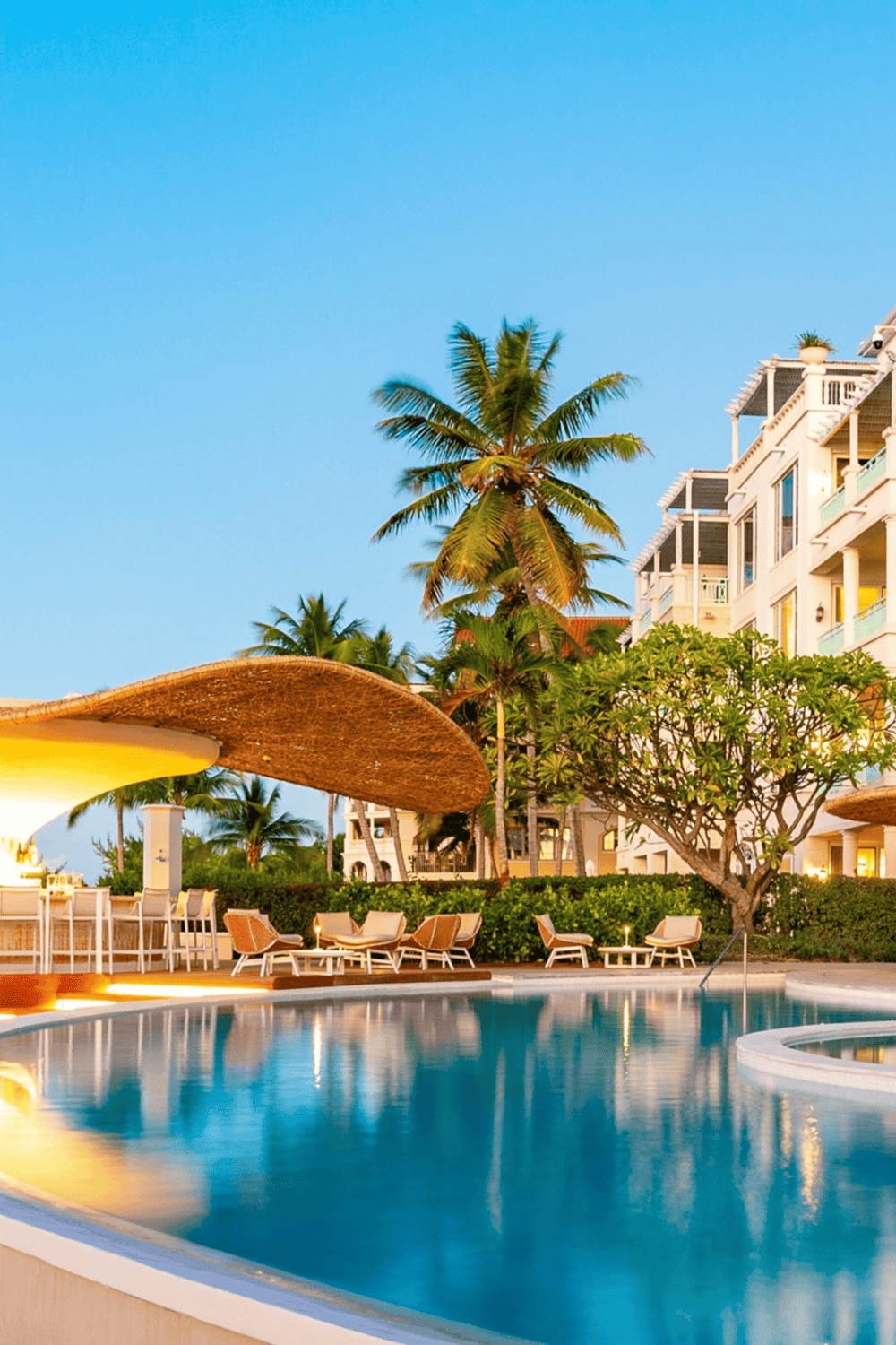 Relaxing hotel pool with palm trees and modern resort buildings at sunset.