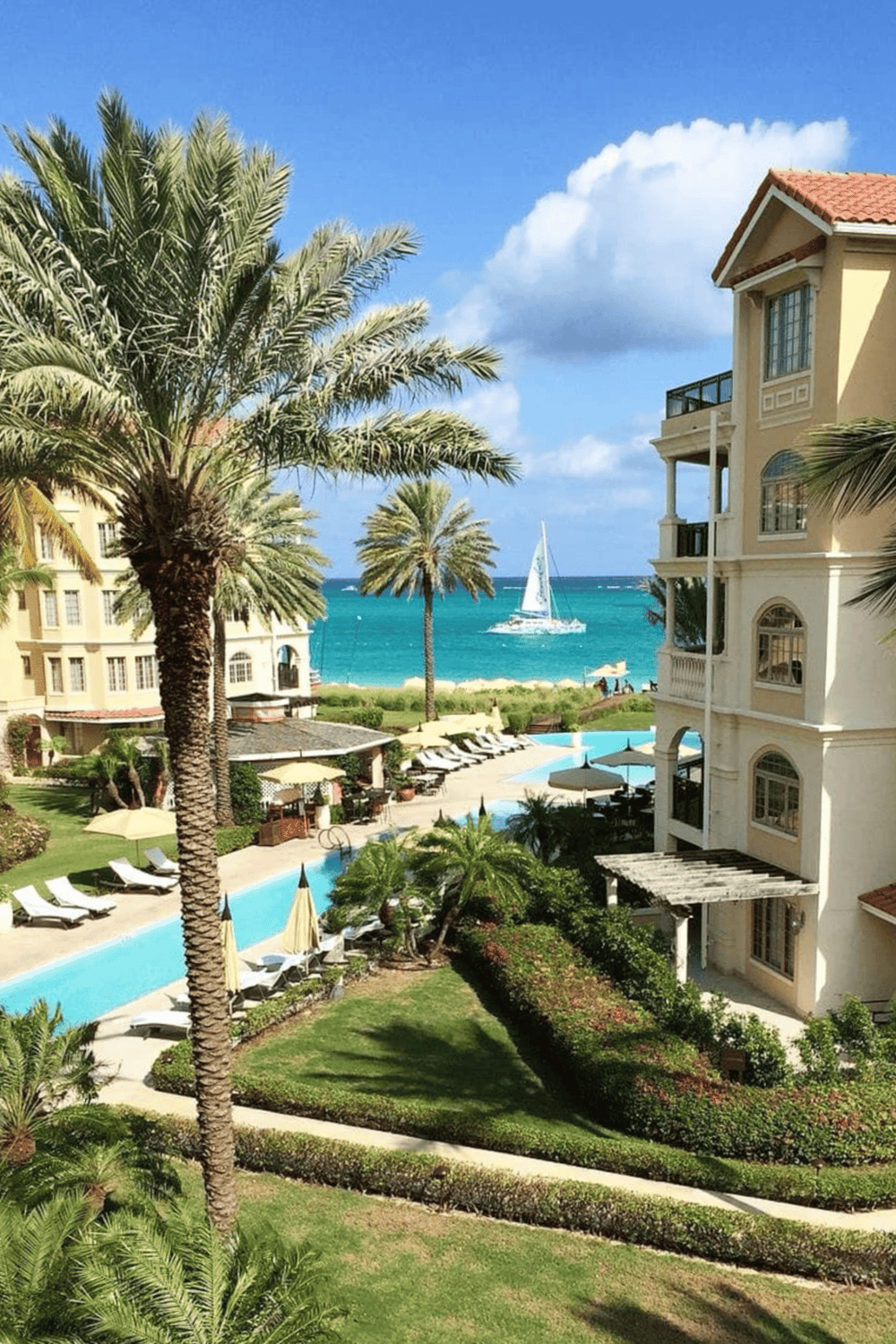 Luxurious beachfront resort with palm trees, swimming pool, ocean view, and sailboat in sunny paradise.