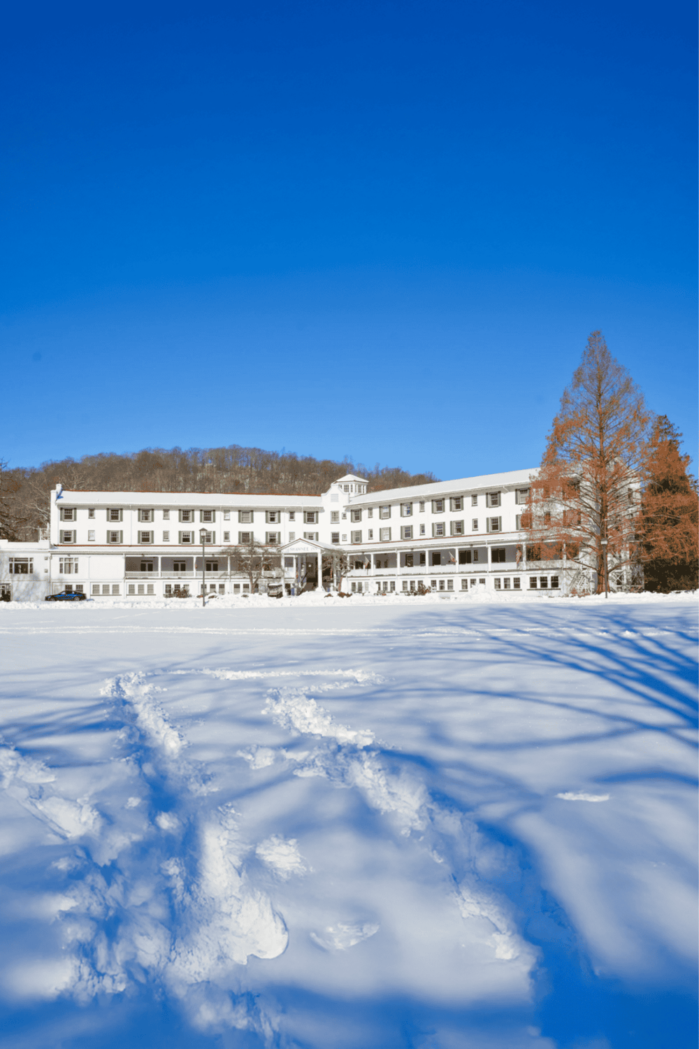 Quiet winter scene of a historic hotel with snow-covered ground, blue sky, and surrounding trees.