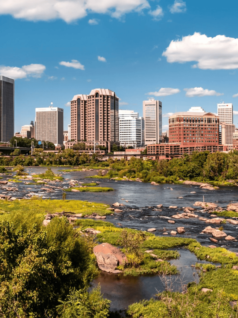 City skyline with tall buildings and a river in foreground under a blue sky with clouds.