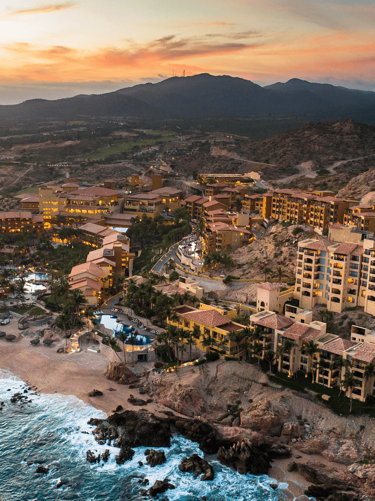 Luxurious seaside resort overlooking rocky beach and ocean with mountain backdrop, sunset sky, and scenic coastal views.
