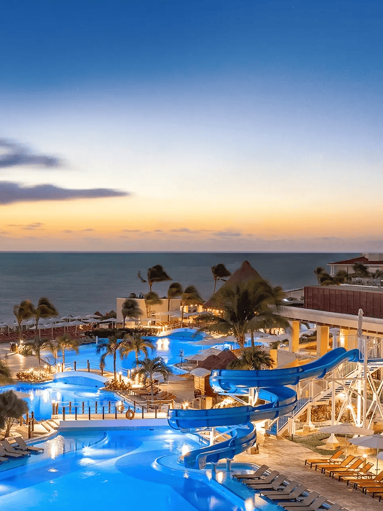 Bright illuminated water slides at a beachfront resort during sunset, showcasing relaxation and adventure.