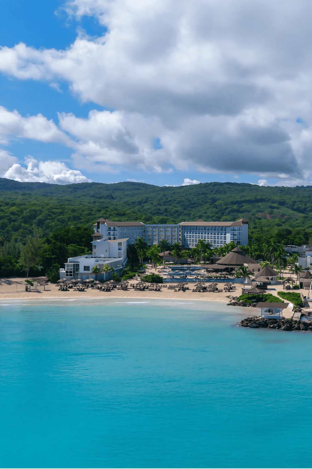 Luxury beachfront resort with ocean view, pool, and lush green hills in the background.