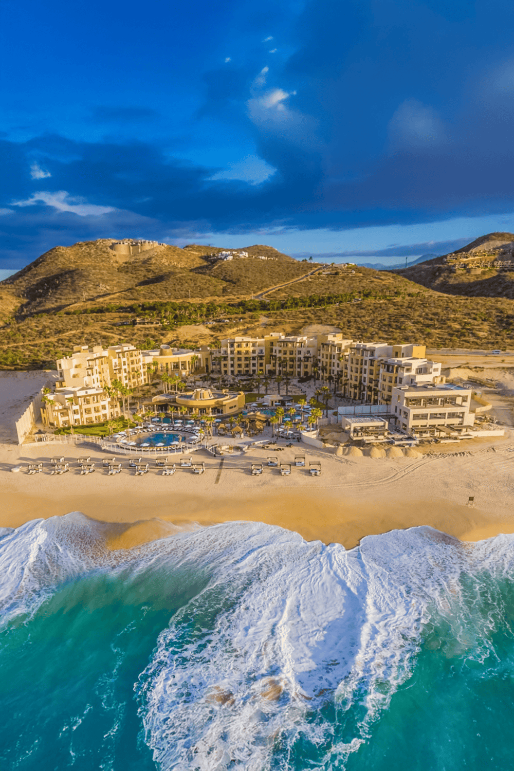Cliffside oceanfront resort with pool, situated on sandy beach, overlooking mountains and blue sky.
