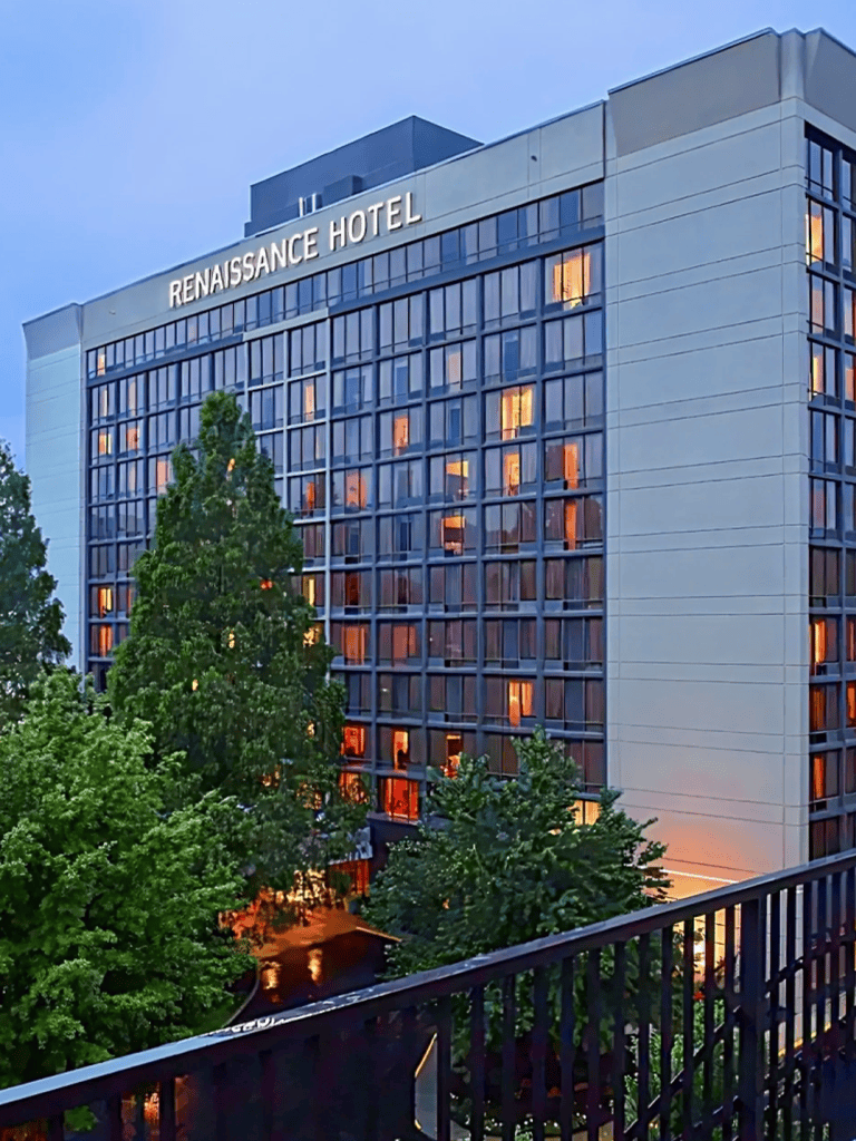 Modern Renaissance Hotel building with illuminated windows at dusk, surrounded by greenery in an urban setting.