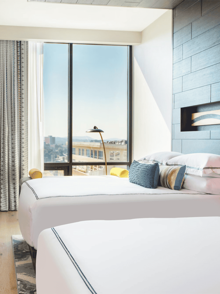 Modern hotel room with large window and city view, stylish interior decor, comfortable bedding, and ambient lighting.