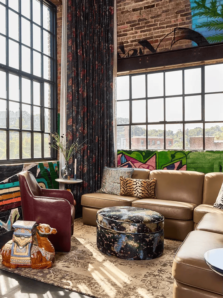 Cozy urban loft with large industrial windows, eclectic furniture, and vibrant wall art, perfect for creative workspace or comfortable lounge.