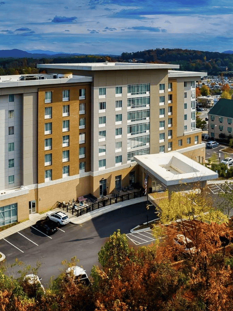 Modern multi-story hotel building with parking lot and scenic mountain view behind.