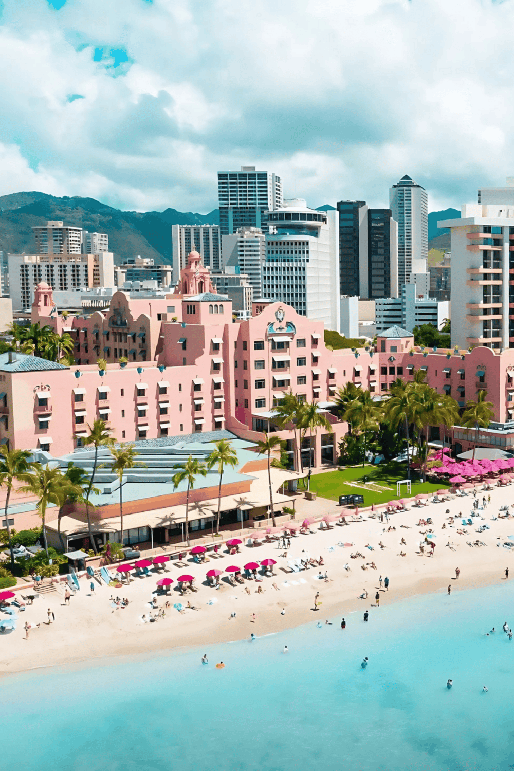 Pink luxury hotel on Waikiki Beach with city skyline and mountains in background, Hawaii travel destination.