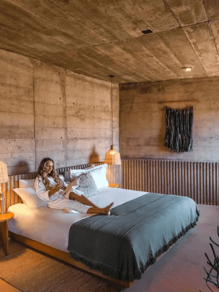 Relaxing woman on cozy bed in modern concrete-walled hotel room with soft lighting.