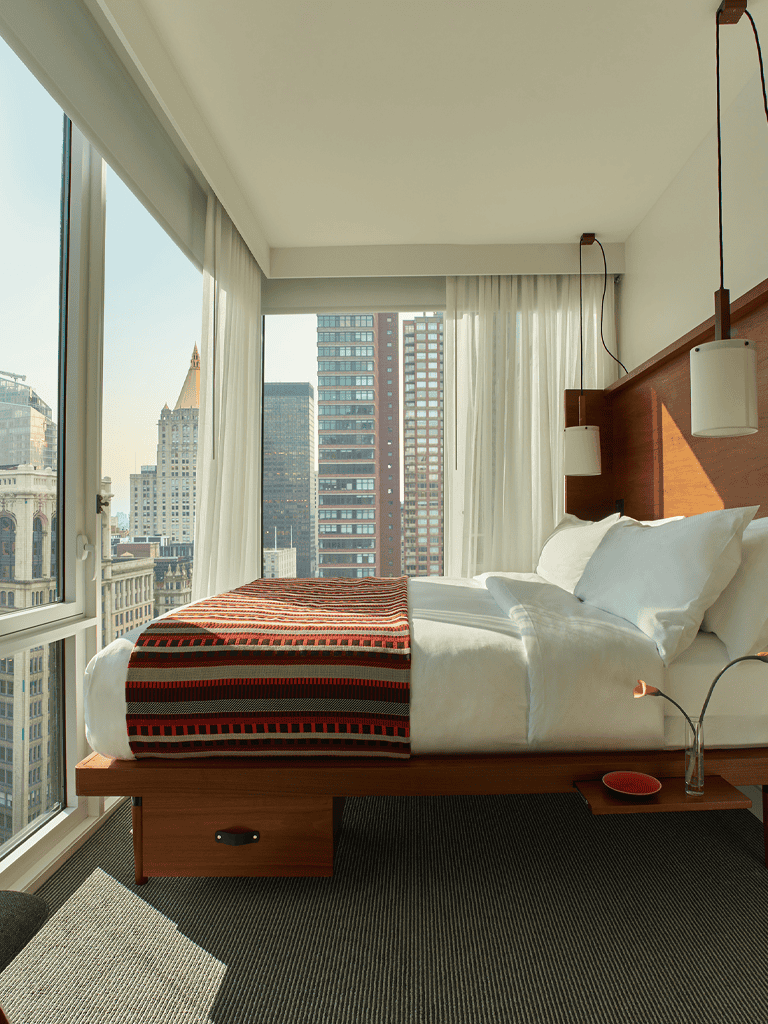 Contemporary hotel room with city view, modern decor, large windows, and comfortable bedding.