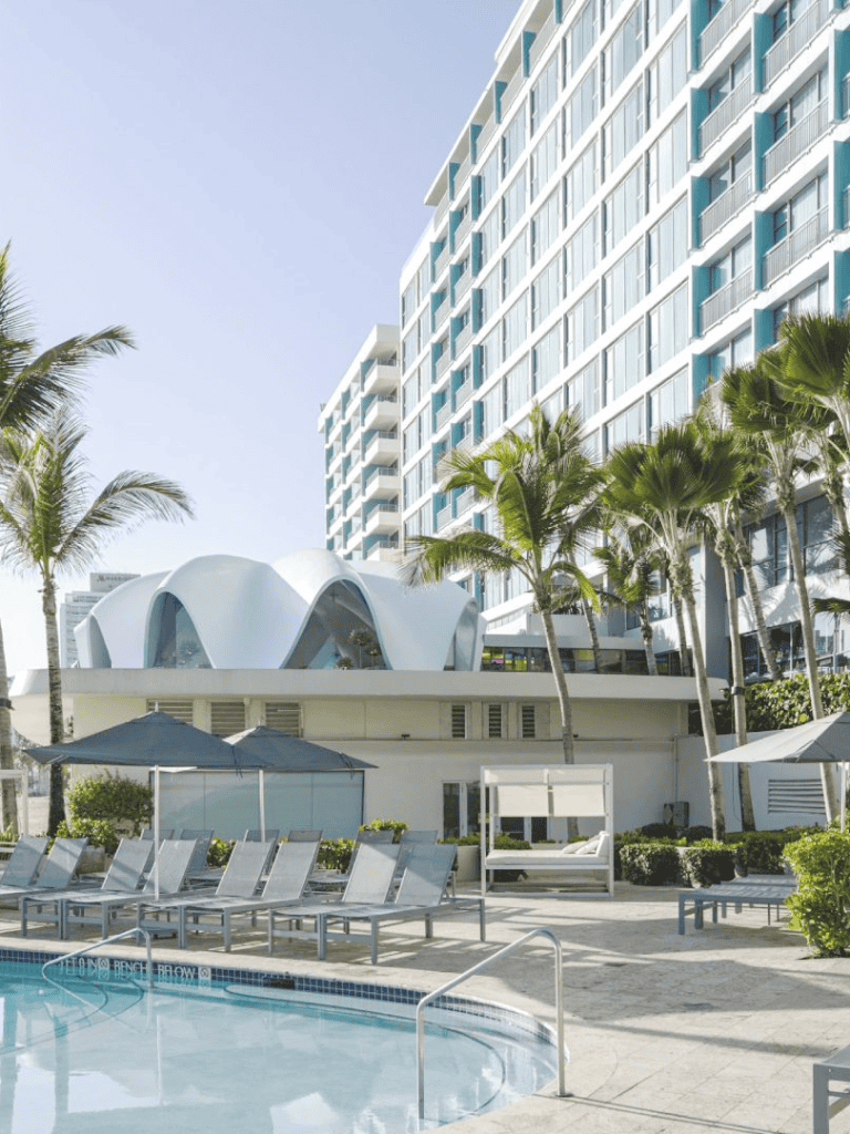 Outdoor hotel pool area with lounge chairs and cabanas in front of a modern building with palm trees.