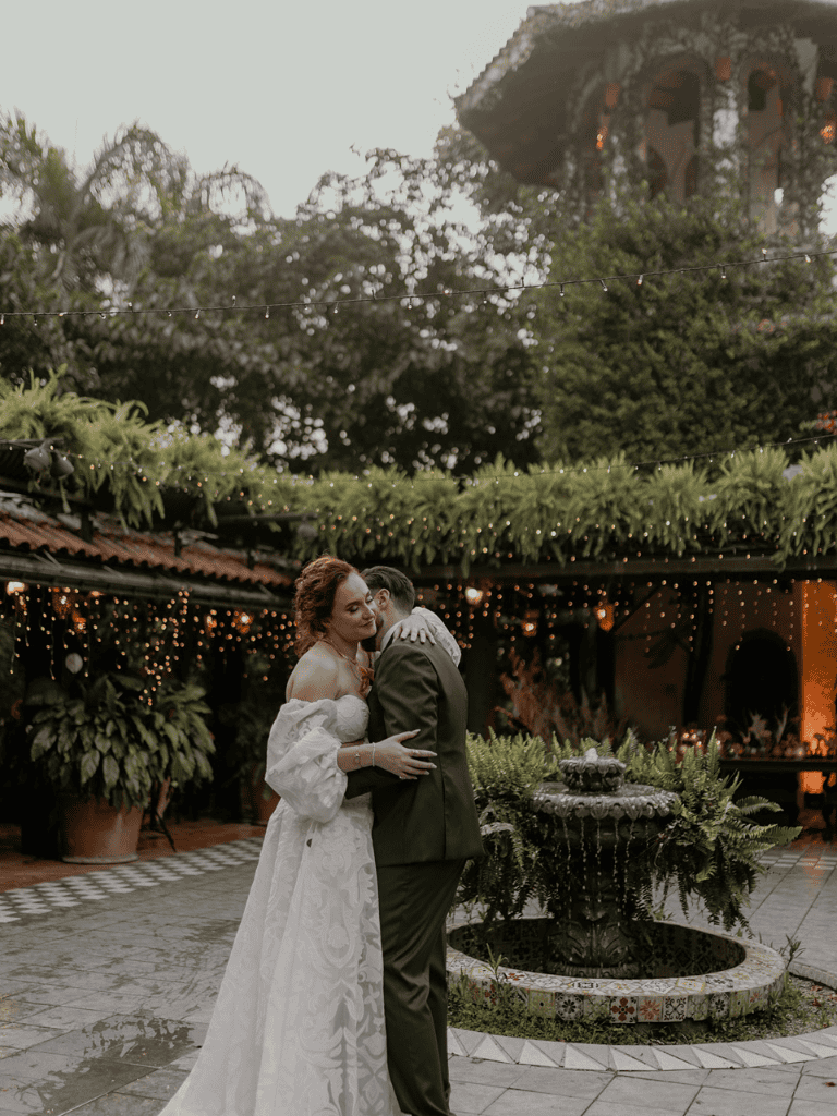 Romantic wedding couple embracing outdoors, lush greenery, fairy lights, and fountain, elegant celebration scene.