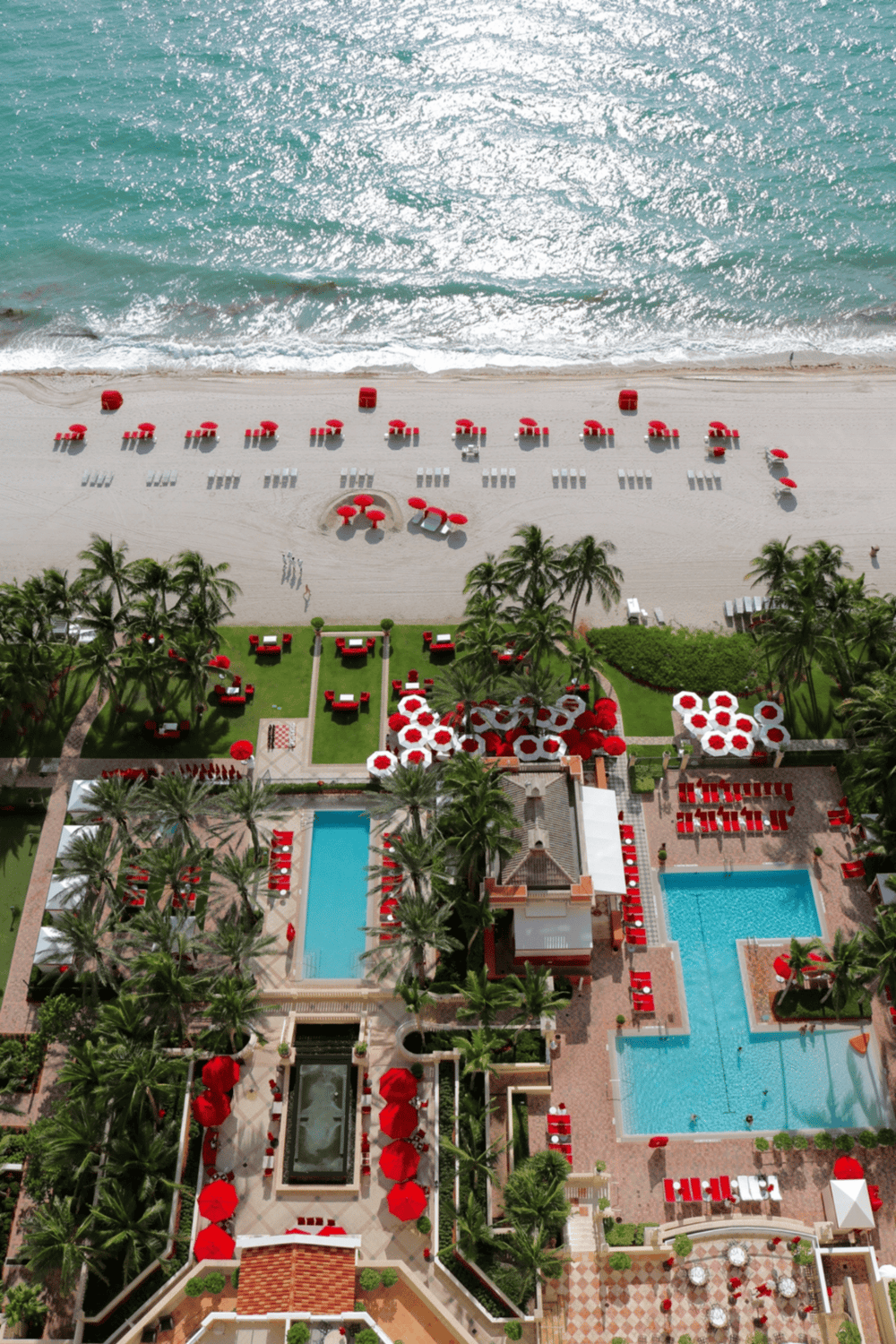 Aerial view of a luxury beachfront resort with pools, palm trees, and beach chairs on sandy shoreline.