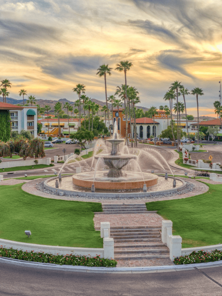 Vibrant fountain in a lush park with palm trees, colorful buildings, and mountain backdrop at sunset.