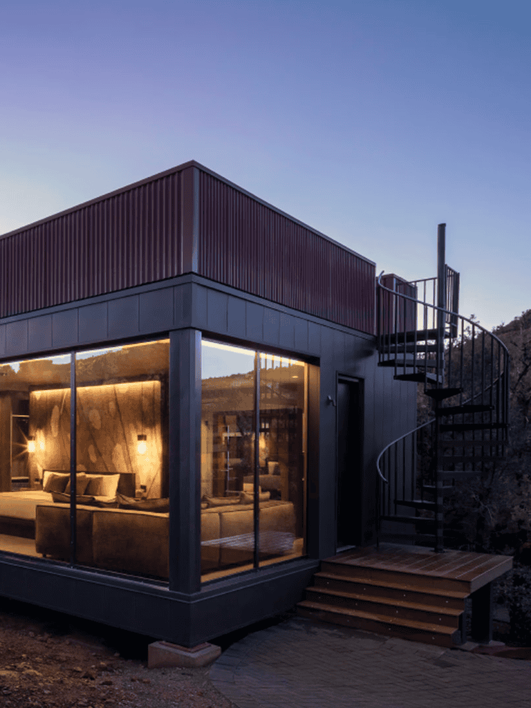 Modern tiny house with large glass windows, outdoor spiral staircase, and scenic mountain view at sunset.