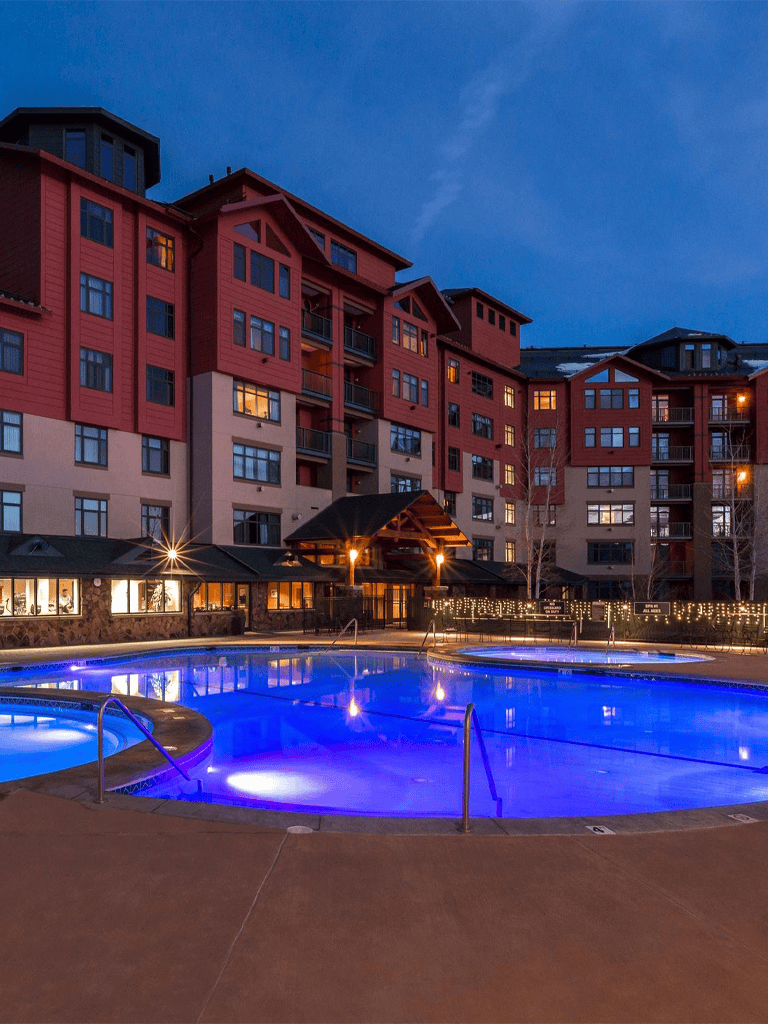 Luxurious hotel pool at dusk with illuminated water and modern apartment building in background.
