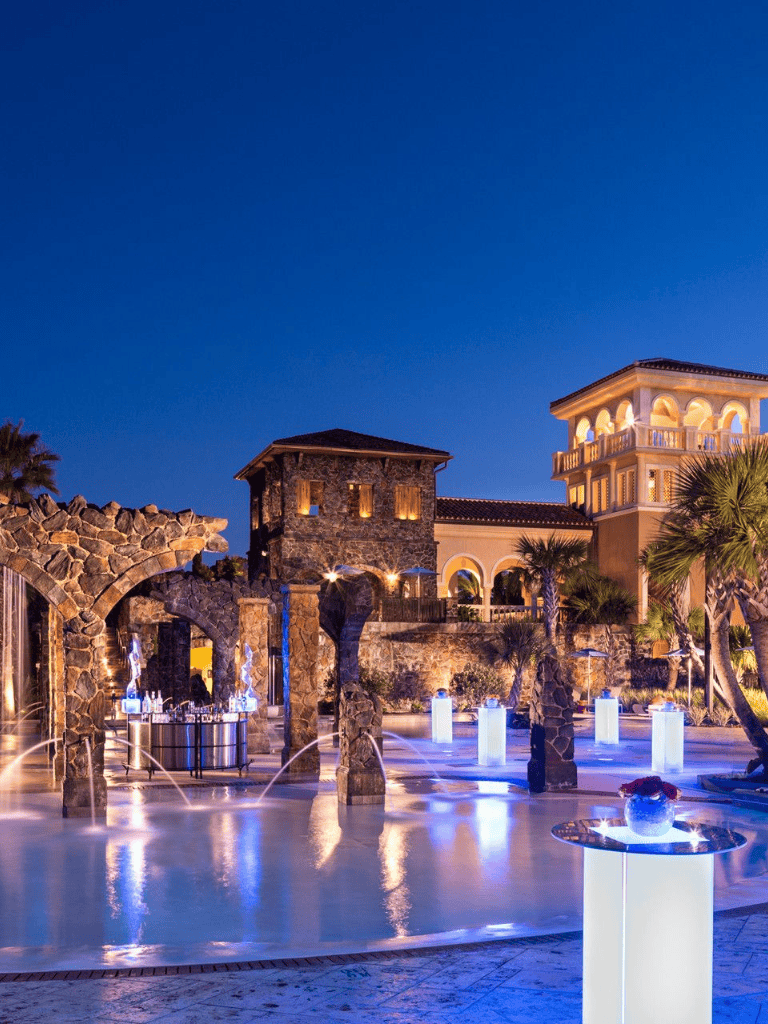 Luxurious outdoor resort pool at night with illuminated fountains and elegant architecture, perfect for vacation relaxation.