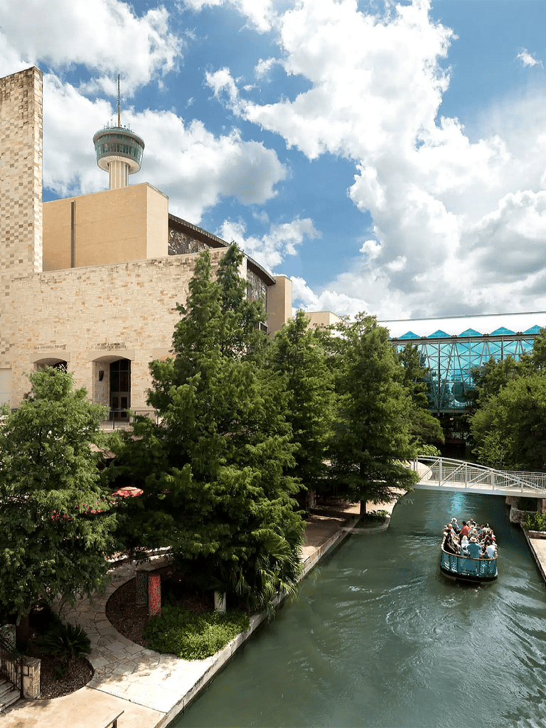 Bright cityscape with a river cruise, lush trees, modern architecture, and city tower under a sunny sky.
