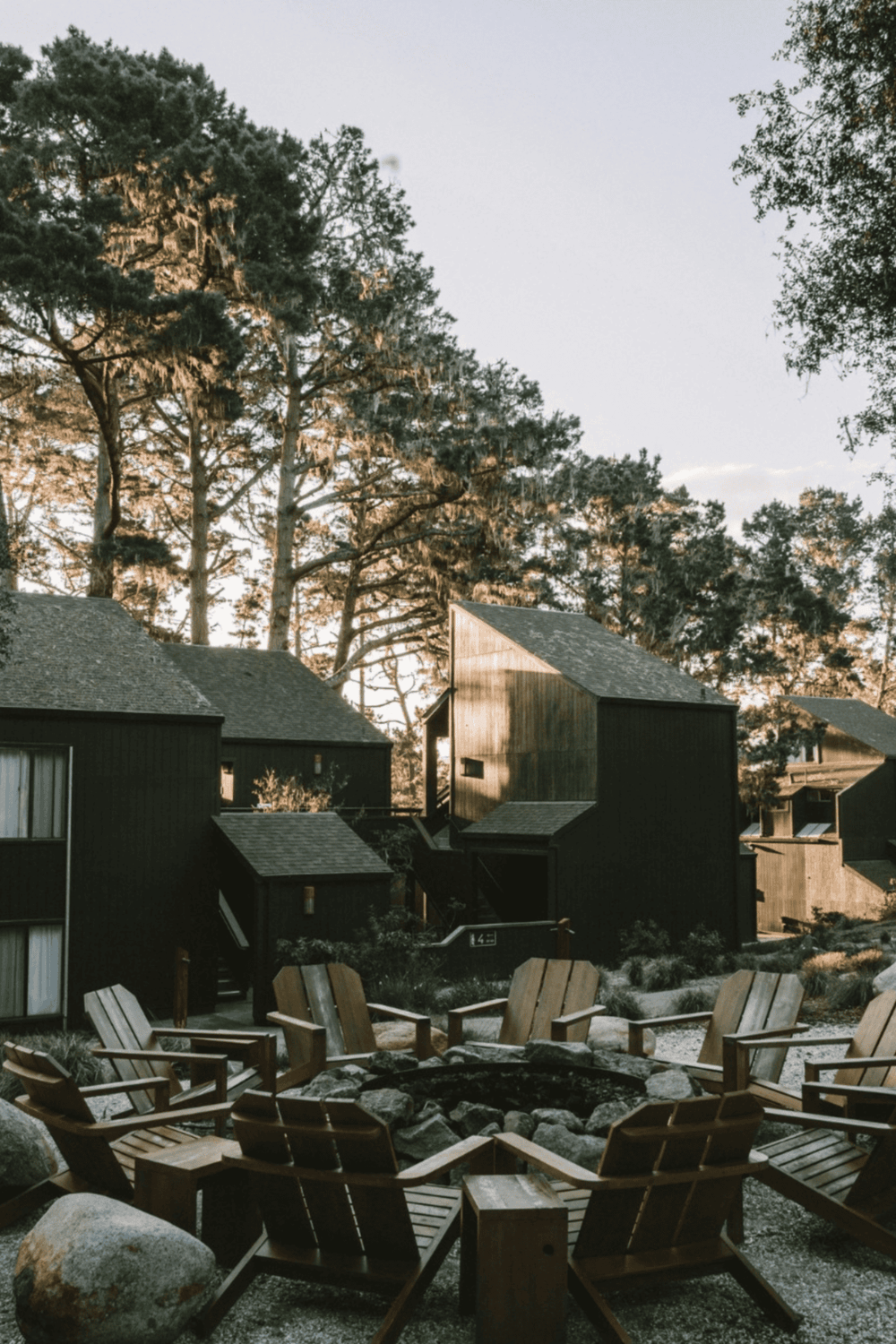 Cozy outdoor fire pit area with Adirondack chairs in front of modern black cabins, surrounded by tall trees at dusk.