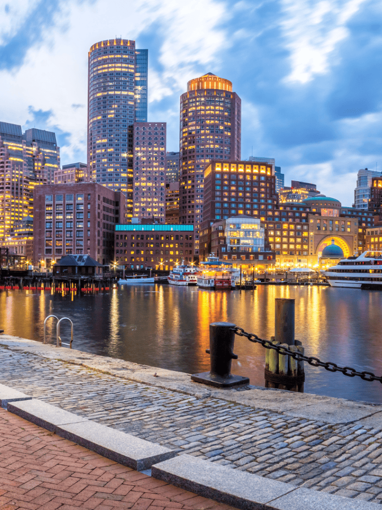 Modern Boston skyline with harbor view at dusk, illuminated skyscrapers and yachts.