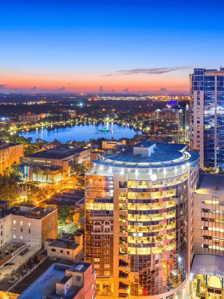 Panoramic city skyline at sunset with water body, modern skyscrapers, and vibrant city lights.
