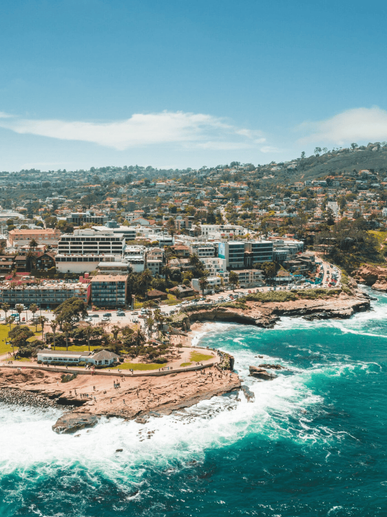 Aerial view of coastal city with buildings and cliffs overlooking the ocean and waves crashing on shore.