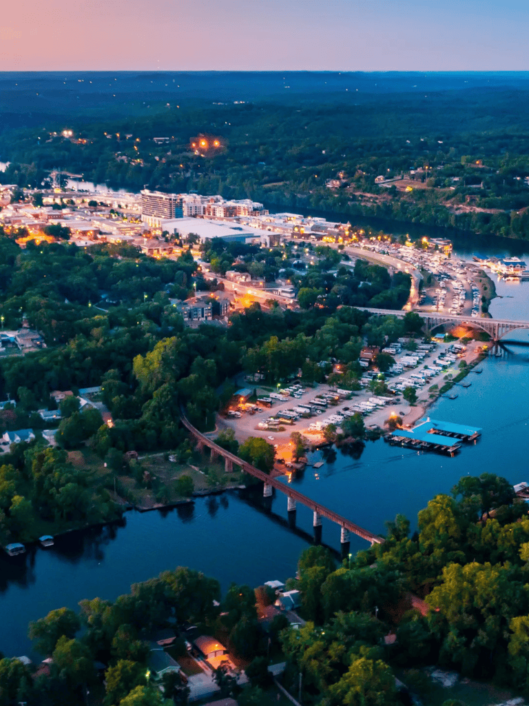 Downtown waterfront at dusk with illuminated buildings and lush green trees.