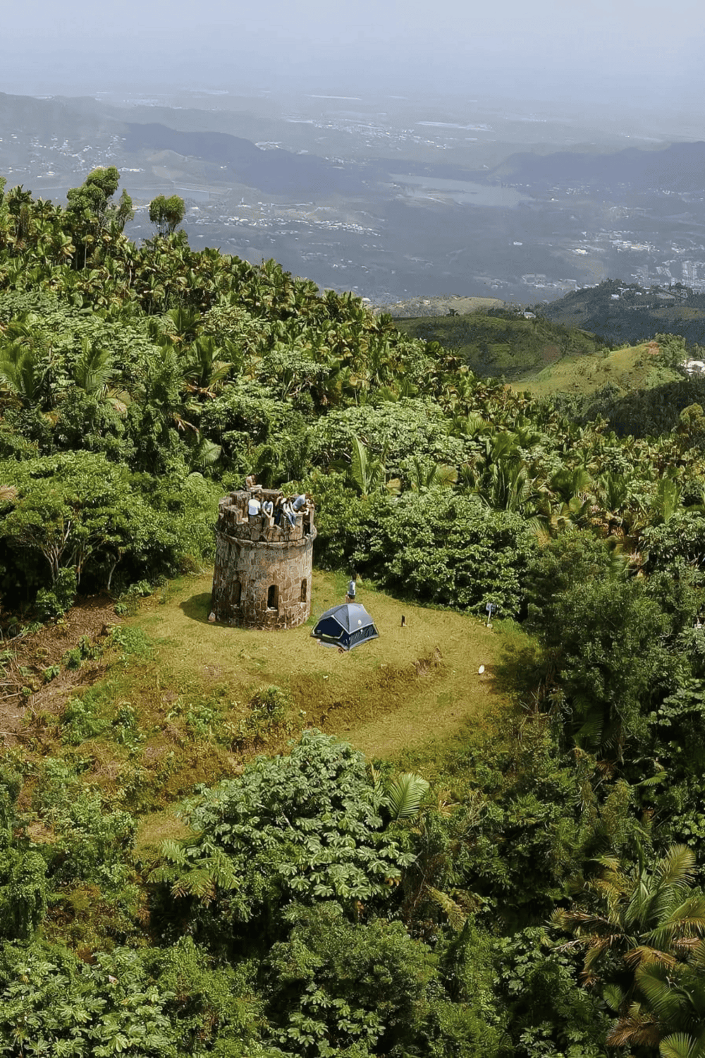 Vibrant green hillside with an old stone tower, camping tent, and lush tropical vegetation in a scenic mountain landscape.