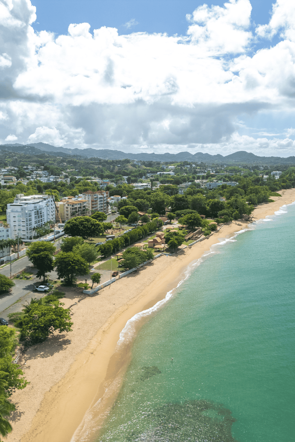 Aerial view of a scenic coastal town with sandy beach, lush greenery, and modern buildings under a partly cloudy sky.