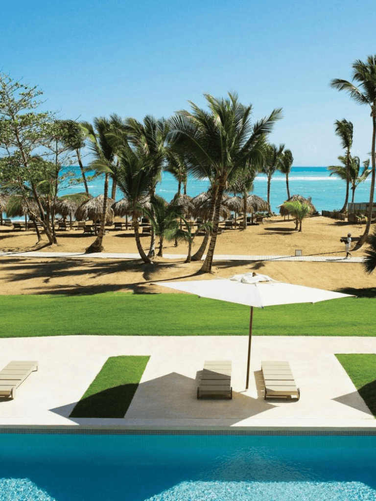 Pool and beach resort with palm trees, lounge chairs, and umbrellas on a sunny day.