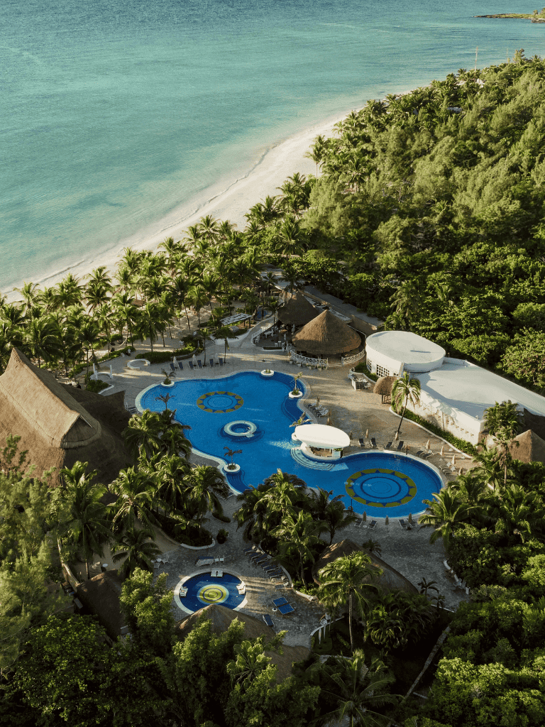 Aerial view of tropical beach resort with pools and lush greenery on a sunny day.