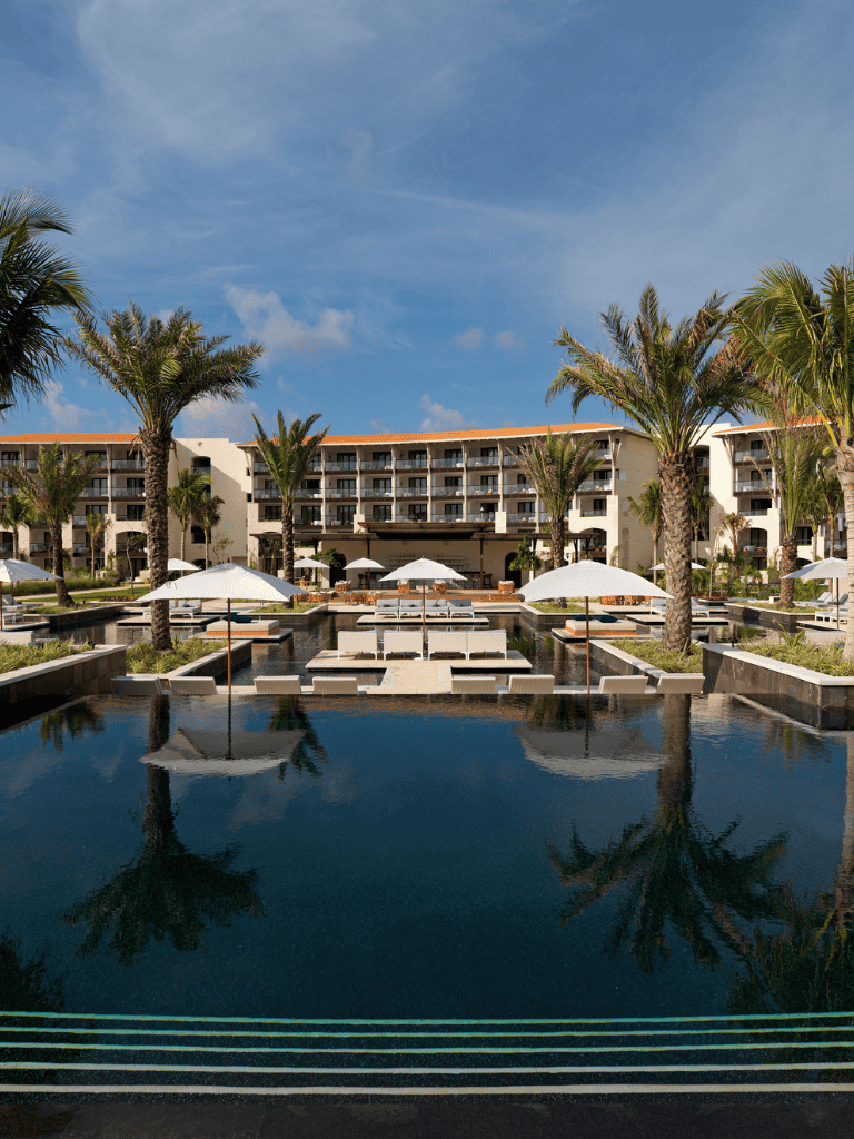 Luxurious hotel pool surrounded by palm trees and modern resort buildings.