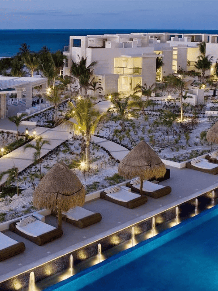 Luxurious beachfront resort with pool, lounge chairs, and palm trees at sunset.