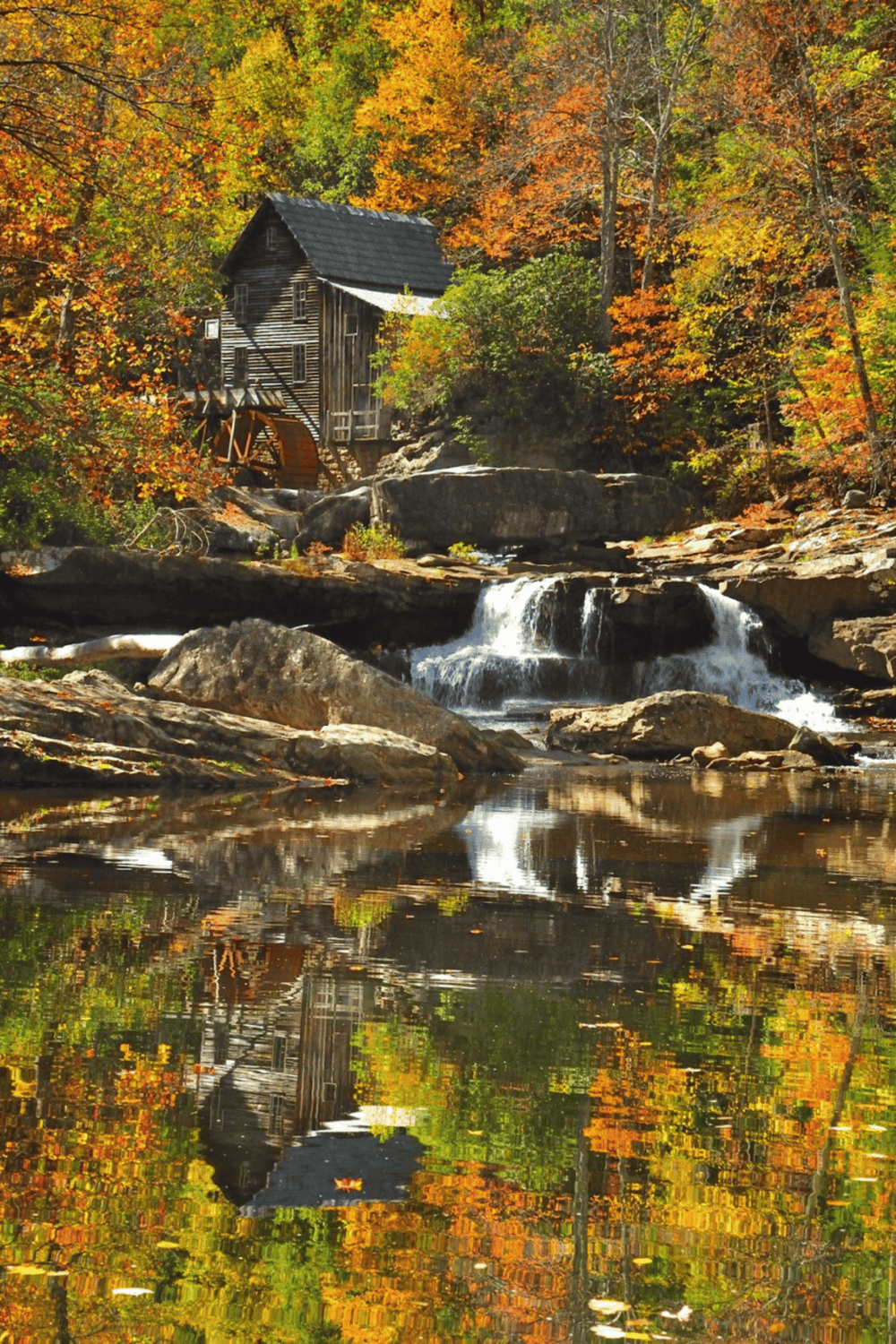 Colorful autumn foliage surrounding a historic wooden watermill and cascading waterfall in a scenic forest setting.