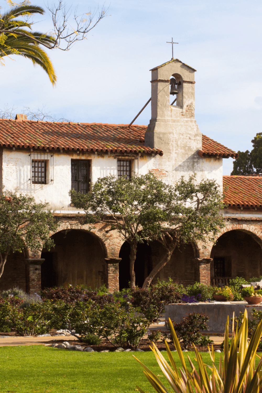 Vivid image of a historic mission-style building with a bell tower, surrounded by lush greenery and colorful plants.