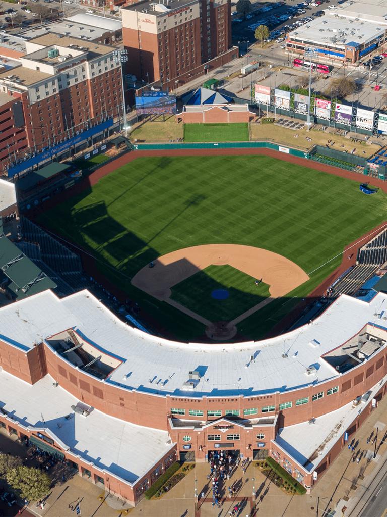 Outdoor baseball stadium with seating, scoreboard, and surrounding cityscape.