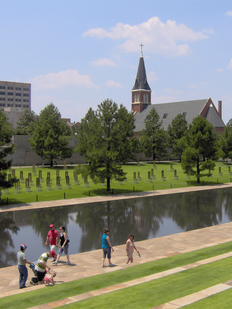 Peaceful park with church and memorials, family walking near water, vibrant greenery, urban skyline background.