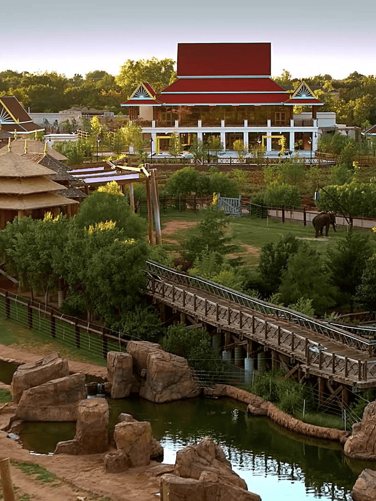Colorful theme park with traditional architecture, lush greenery, rides, and a small zoo area with an elephant.